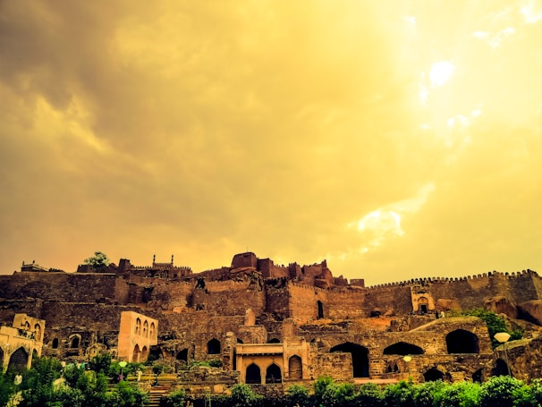 A sprawling, ancient fortress sits atop a hill, characterized by its stone walls and towers. The sky is painted a dramatic golden-yellow, suggesting sunset or sunrise, casting a warm glow over the landscape. Dense greenery surrounds the base of the fortress.