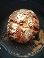 A rustic round loaf of bread with a golden-brown crust, dusted with flour and sitting on a dark baking sheet. The surface of the bread is cracked, giving it a homemade, artisanal appearance.