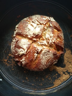 A rustic round loaf of bread with a golden-brown crust, dusted with flour and sitting on a dark baking sheet. The surface of the bread is cracked, giving it a homemade, artisanal appearance.