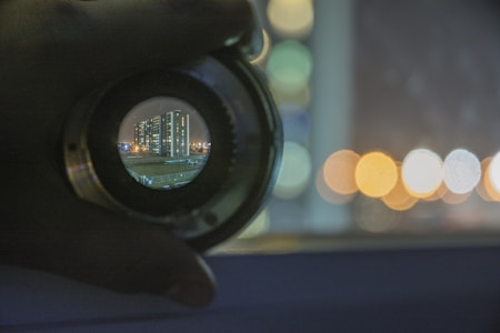 A hand holds a camera lens, through which a cityscape with tall buildings is visible. The background features blurred bokeh lights, suggesting a nighttime or dusk setting.