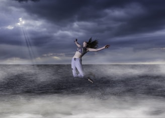 A person is leaping joyfully above the ocean, surrounded by misty waves. The sky is overcast with dramatic dark clouds, and a few rays of light break through, creating a dynamic and striking atmosphere.