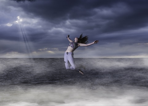 A person is leaping joyfully above the ocean, surrounded by misty waves. The sky is overcast with dramatic dark clouds, and a few rays of light break through, creating a dynamic and striking atmosphere.