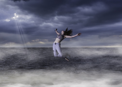 A person is leaping joyfully above the ocean, surrounded by misty waves. The sky is overcast with dramatic dark clouds, and a few rays of light break through, creating a dynamic and striking atmosphere.