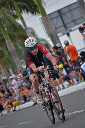 A cyclist wearing a helmet and racing attire is actively participating in a competitive cycling event. The scene is lively, with spectators lined up along the sides of the road, some sitting and others standing, all observing the race. Palm trees are visible in the background, suggesting a warm climate.