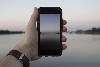 A hand holding a smartphone against a serene, blurred landscape. The phone displays a tranquil scene of a calm body of water and distant land against a gentle evening or morning sky.