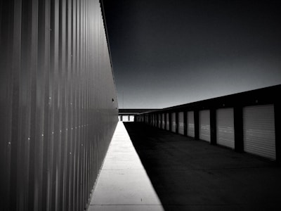 A series of storage units with roll-up doors extends into the distance. The perspective view is along the side of a large metal building, casting long shadows on the ground. The overall atmosphere is empty and quiet.