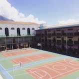 A school courtyard with a large multi-sport area featuring basketball and volleyball courts. The building surrounds the court on three sides and has multiple stories with balconies. A mountain and a partly cloudy sky are visible in the background.
