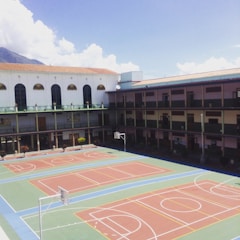 A school courtyard with a large multi-sport area featuring basketball and volleyball courts. The building surrounds the court on three sides and has multiple stories with balconies. A mountain and a partly cloudy sky are visible in the background.