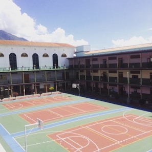 A school courtyard with a large multi-sport area featuring basketball and volleyball courts. The building surrounds the court on three sides and has multiple stories with balconies. A mountain and a partly cloudy sky are visible in the background.