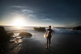 A lone surfer stands on a sandy beach holding a surfboard under their arm, facing the ocean as the sun sets over the horizon. The waves crash gently against the rocks nearby, creating a serene and contemplative atmosphere.