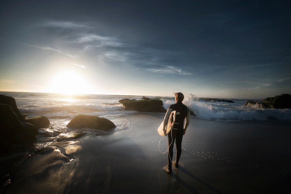 man standing carrying surfboard near seashore during daytime