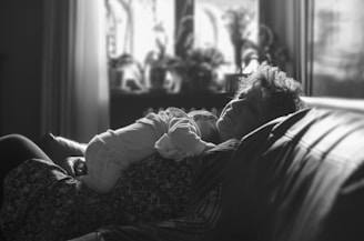 A black and white image of a person reclining on a couch with a young child resting on their chest. Natural light filters through the window in the background, softly illuminating the scene. The setting features potted plants on a windowsill.