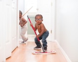 Two young children are playfully cleaning a hallway with mops. One child, standing closer to the camera, is holding a red mop, wearing glasses, jeans, and a necklace. The other child is in the background, wearing suspenders and holding a broom.