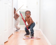Two young children are playfully cleaning a hallway with mops. One child, standing closer to the camera, is holding a red mop, wearing glasses, jeans, and a necklace. The other child is in the background, wearing suspenders and holding a broom.