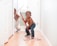 Two young children are playfully cleaning a hallway with mops. One child, standing closer to the camera, is holding a red mop, wearing glasses, jeans, and a necklace. The other child is in the background, wearing suspenders and holding a broom.