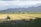 A vast agricultural field with a tractor working on the land. Rows of freshly plowed earth stretch across the horizon, bordered by a line of trees and distant mountains under a partly cloudy sky.