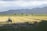 A vast agricultural field with a tractor working on the land. Rows of freshly plowed earth stretch across the horizon, bordered by a line of trees and distant mountains under a partly cloudy sky.