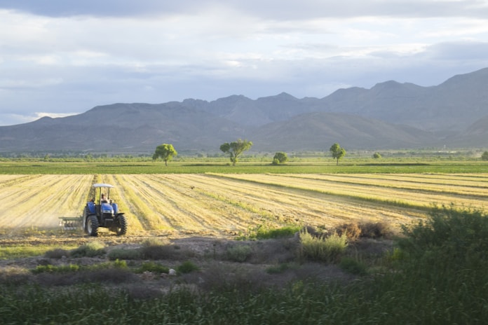 A vast agricultural field with a tractor working on the land. Rows of freshly plowed earth stretch across the horizon, bordered by a line of trees and distant mountains under a partly cloudy sky.