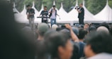 A large outdoor crowd gathered for a concert with a band playing in the background. Musicians are performing on stage with instruments like a guitar and keyboard, while a cameraman captures the event.