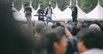 A large outdoor crowd gathered for a concert with a band playing in the background. Musicians are performing on stage with instruments like a guitar and keyboard, while a cameraman captures the event.