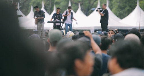 A large outdoor crowd gathered for a concert with a band playing in the background. Musicians are performing on stage with instruments like a guitar and keyboard, while a cameraman captures the event.