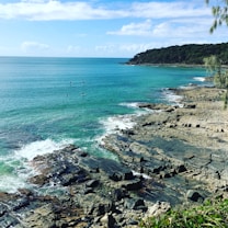 A coastal landscape with clear turquoise waters meeting rocky shores. In the background, a tree-covered hill partially obscures the horizon under a partly cloudy sky. Three paddleboarders are visible in the water, adding a human element to the serene setting.