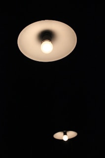 Two ceiling light fixtures are illuminated against a dark background. The closer fixture is larger and more prominent, casting a gentle glow.