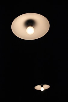 Two ceiling light fixtures are illuminated against a dark background. The closer fixture is larger and more prominent, casting a gentle glow.