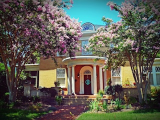 A charming two-story brick house surrounded by lush greenery and blooming pink and white flowers. The entrance features a grand arched doorway with decorative columns. Tall trees with abundant blossoms frame the property, creating a picturesque and inviting atmosphere.