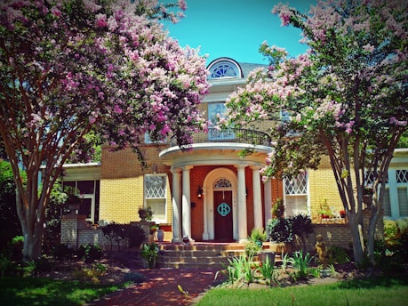A charming two-story brick house surrounded by lush greenery and blooming pink and white flowers. The entrance features a grand arched doorway with decorative columns. Tall trees with abundant blossoms frame the property, creating a picturesque and inviting atmosphere.