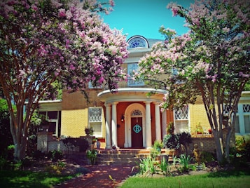 A charming two-story brick house surrounded by lush greenery and blooming pink and white flowers. The entrance features a grand arched doorway with decorative columns. Tall trees with abundant blossoms frame the property, creating a picturesque and inviting atmosphere.