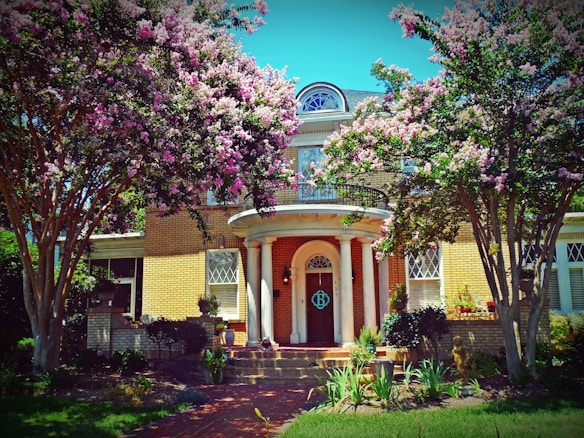 A charming two-story brick house surrounded by lush greenery and blooming pink and white flowers. The entrance features a grand arched doorway with decorative columns. Tall trees with abundant blossoms frame the property, creating a picturesque and inviting atmosphere.