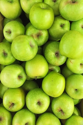 A pile of bright green apples closely packed together, showcasing their smooth skin and fresh appearance. The apples vary slightly in shade, with some displaying a hint of yellow or greenish-brown spots near the stem.