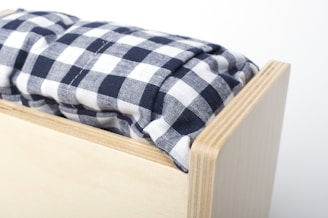 A close-up view of a wooden box with a soft cushion on top. The cushion features a black and white checkered pattern, creating a cozy and homely feel. The wood has a light finish with visible grain, suggesting simplicity and functionality.
