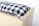 A close-up view of a wooden box with a soft cushion on top. The cushion features a black and white checkered pattern, creating a cozy and homely feel. The wood has a light finish with visible grain, suggesting simplicity and functionality.