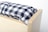 A close-up view of a wooden box with a soft cushion on top. The cushion features a black and white checkered pattern, creating a cozy and homely feel. The wood has a light finish with visible grain, suggesting simplicity and functionality.