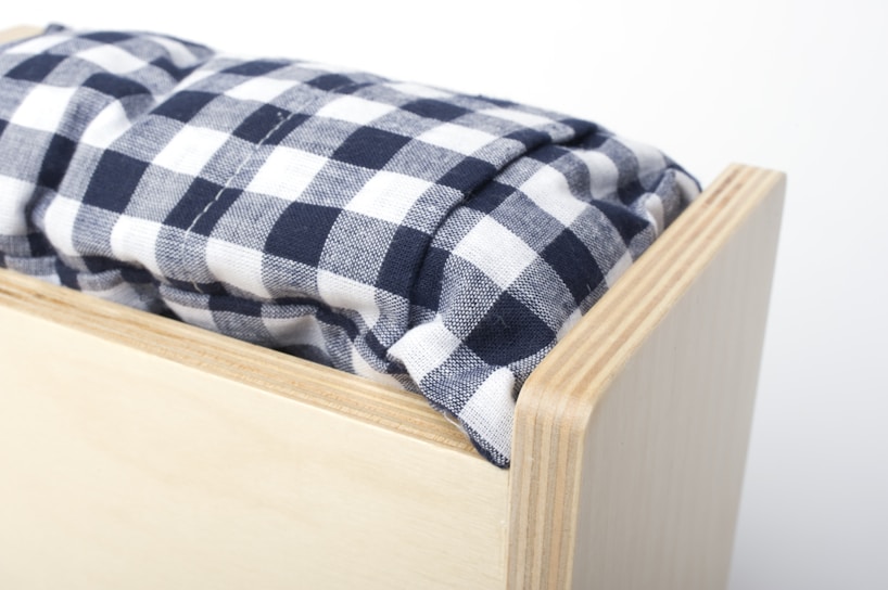 A close-up view of a wooden box with a soft cushion on top. The cushion features a black and white checkered pattern, creating a cozy and homely feel. The wood has a light finish with visible grain, suggesting simplicity and functionality.