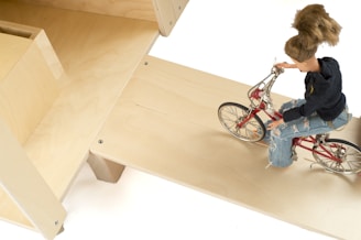 A doll with detailed features is riding a small red bicycle. The doll is dressed in a black hoodie and ripped blue jeans. The setting includes light-colored wooden structures that form ramps or platforms.