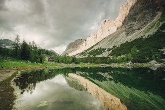 A tranquil mountain landscape featuring a reflective lake surrounded by lush green forests and steep rocky cliffs. A small wooden cabin is nestled among the trees, creating a sense of seclusion and harmony with nature. The sky is overcast, adding a dramatic contrast to the vibrant greenery and the serene water.