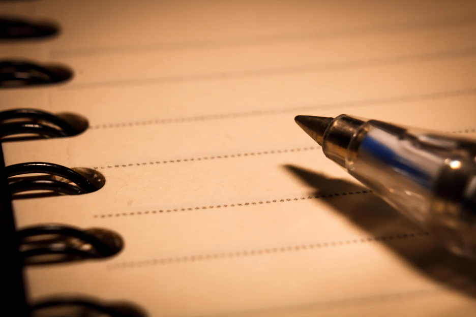 Close-up of a sleek metal ballpoint pen resting on a wooden desk beside an open notebook.