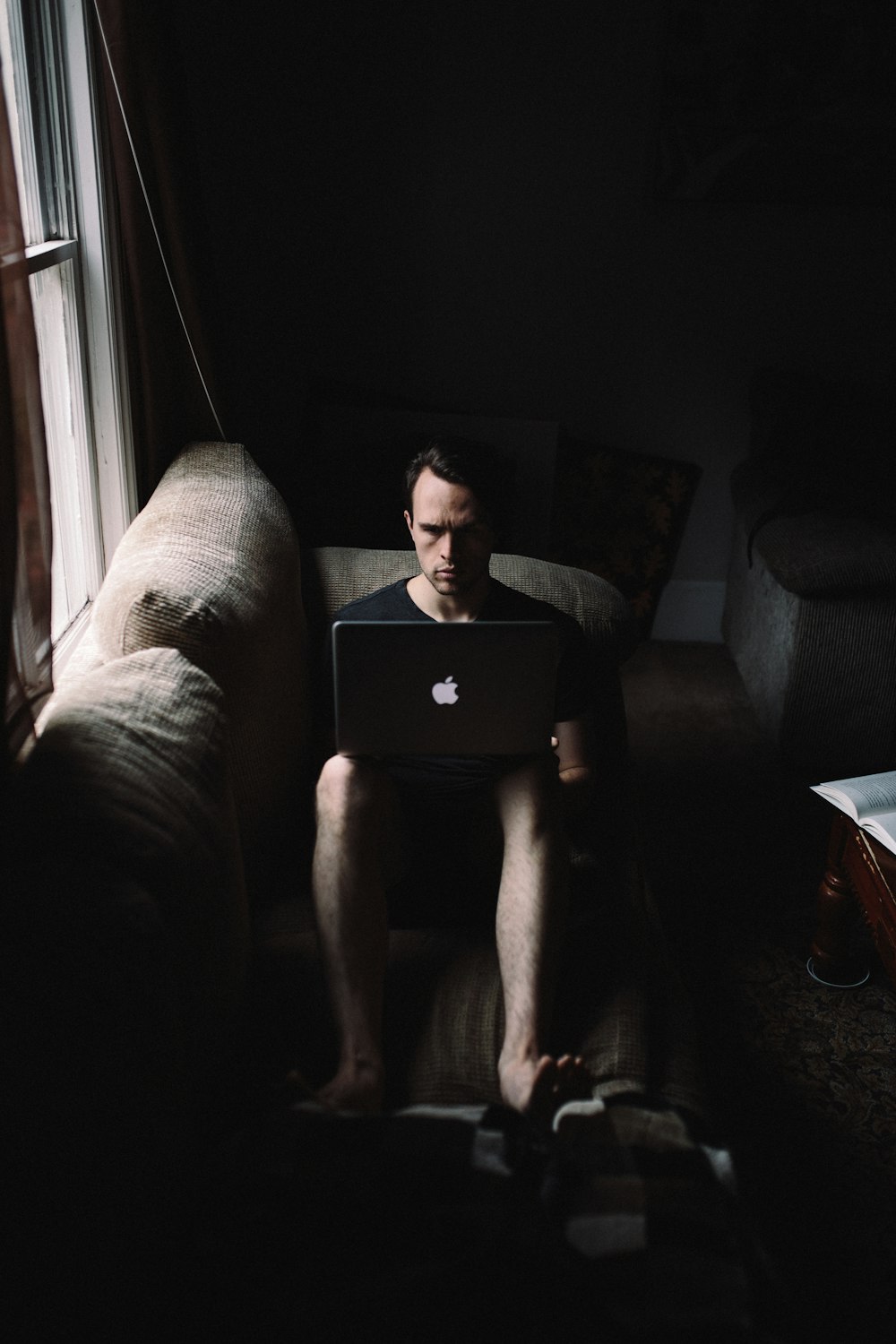 창문 옆에서 MacBook을 사용하여 소파에 앉아있는 남자