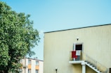 A modern residential building with a bright yellow exterior features a staircase leading to a slightly open door with red and white towels hanging on the railing. A lush green tree is visible in the foreground, and additional buildings can be seen in the background under a clear blue sky.