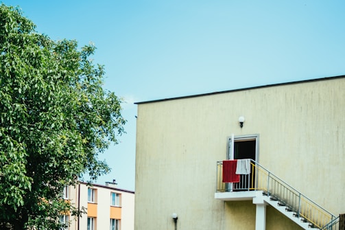 A modern residential building with a bright yellow exterior features a staircase leading to a slightly open door with red and white towels hanging on the railing. A lush green tree is visible in the foreground, and additional buildings can be seen in the background under a clear blue sky.
