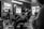 A black-and-white image of a barbershop setting with a focus on a statue in the foreground. The barber and client are slightly out of focus, with the client seated in a barber's chair. The room is filled with ambient lighting, and there are multiple reflections visible in mirrors.