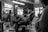 A black-and-white image of a barbershop setting with a focus on a statue in the foreground. The barber and client are slightly out of focus, with the client seated in a barber's chair. The room is filled with ambient lighting, and there are multiple reflections visible in mirrors.