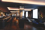 A modern, dimly-lit bar with a sleek, sophisticated interior. It features a long counter with bar stools, and a series of cozy seating areas with plush couches and small round tables. The space is elegantly decorated with dark tones and reflective surfaces, alongside large windows allowing natural light to enter.