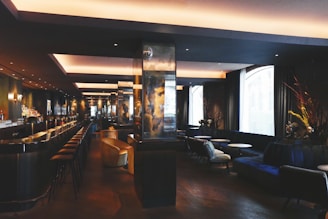 A modern, dimly-lit bar with a sleek, sophisticated interior. It features a long counter with bar stools, and a series of cozy seating areas with plush couches and small round tables. The space is elegantly decorated with dark tones and reflective surfaces, alongside large windows allowing natural light to enter.