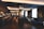 A modern, dimly-lit bar with a sleek, sophisticated interior. It features a long counter with bar stools, and a series of cozy seating areas with plush couches and small round tables. The space is elegantly decorated with dark tones and reflective surfaces, alongside large windows allowing natural light to enter.