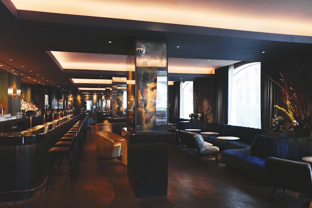 A modern, dimly-lit bar with a sleek, sophisticated interior. It features a long counter with bar stools, and a series of cozy seating areas with plush couches and small round tables. The space is elegantly decorated with dark tones and reflective surfaces, alongside large windows allowing natural light to enter.