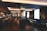 A modern, dimly-lit bar with a sleek, sophisticated interior. It features a long counter with bar stools, and a series of cozy seating areas with plush couches and small round tables. The space is elegantly decorated with dark tones and reflective surfaces, alongside large windows allowing natural light to enter.